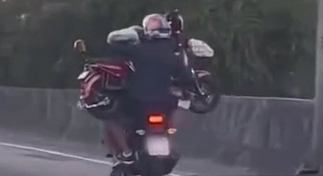Homem pilota moto enquanto garupa carrega outro veículo